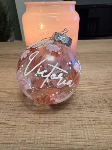 A decorative glass ornament named "Victoria" with pink floral design beside a candle.